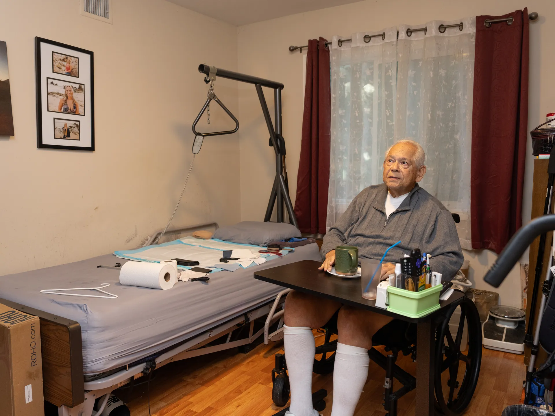 Resident in wheelchair at accessible bedroom table with hospital bed and mobility equipment visible